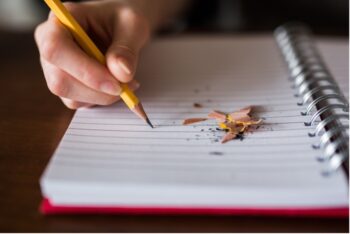 Child's hand writing on a notebook with pencil shavings on the page